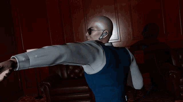 a bald man wearing sunglasses and a blue vest is standing in front of a couch