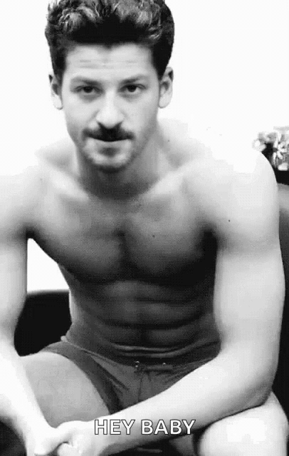 a shirtless man with a mustache is sitting on a couch with his hands on his knees .