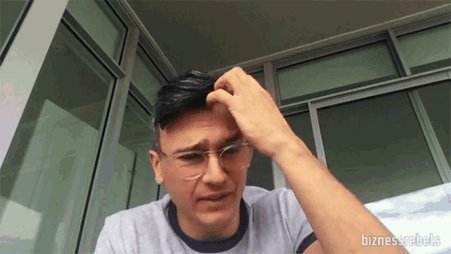 a man wearing glasses scratches his head with his hand in front of a building with the words bizness rebels below him