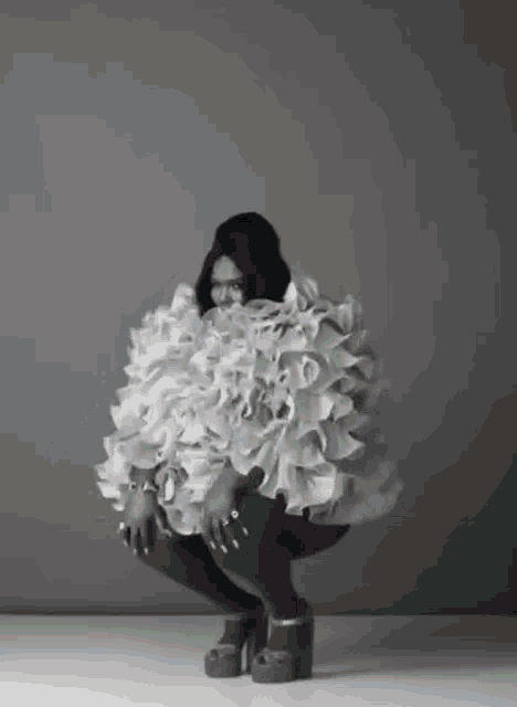 a black and white photo of a woman wearing a ruffled dress and heels squatting down .