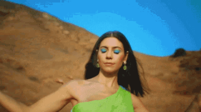 a woman in a green dress and blue eye shadow is standing in the desert with her eyes closed .