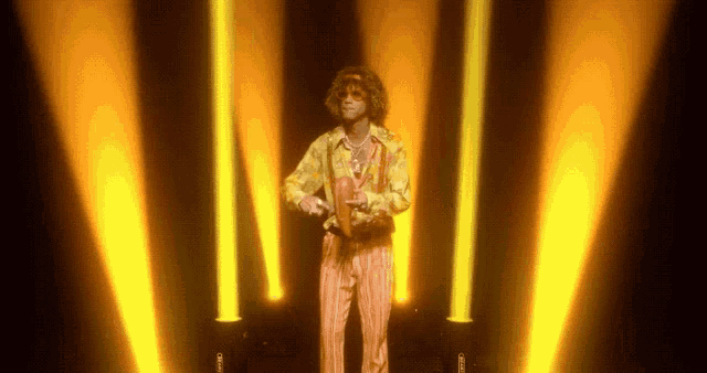 a man in a yellow shirt and orange pants is standing in front of a stage with yellow lights behind him