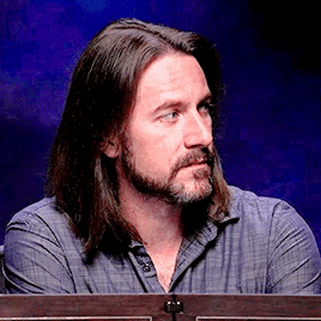 a man with long hair and a beard sits in front of a blue background