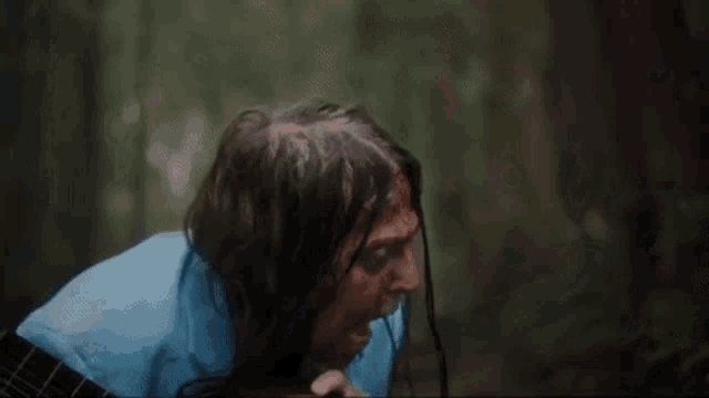 a man with long hair is standing in the woods with blood on his face .