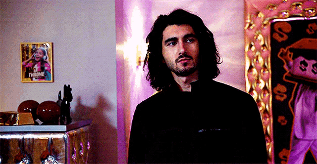 a man with long hair and a beard is standing in front of a pink wall with a picture on it that says " throne "