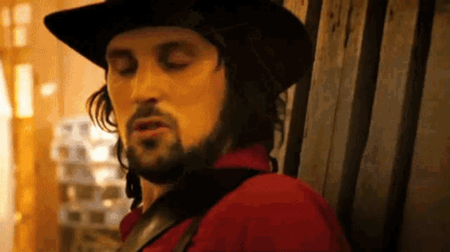 a man with a beard wearing a cowboy hat and a red shirt is leaning against a wooden wall .