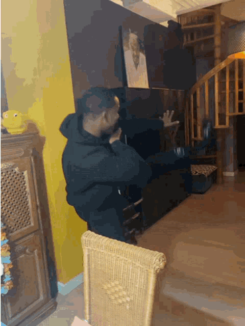 a man in a black hoodie sits in a living room with a wicker chair