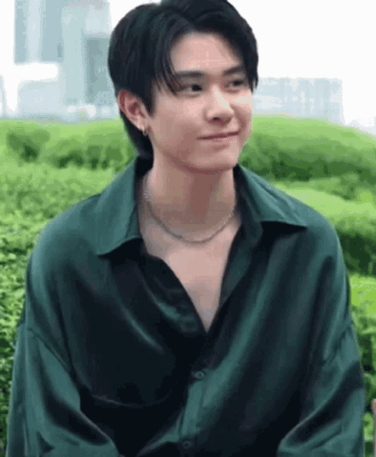 a young man wearing a green shirt and a necklace is smiling while sitting in front of a bush .