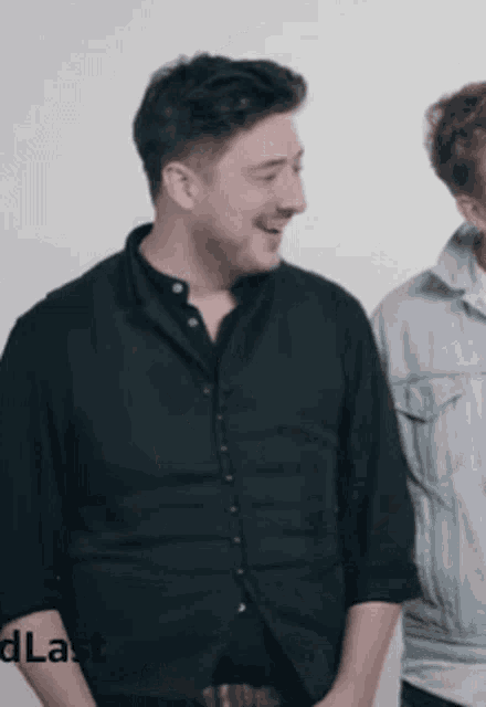 a man in a black shirt is laughing while standing next to another man in a denim jacket