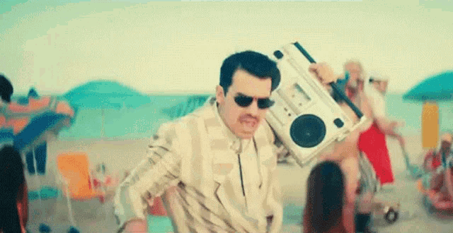 a man in a suit and sunglasses is holding a boombox on his shoulder on the beach .