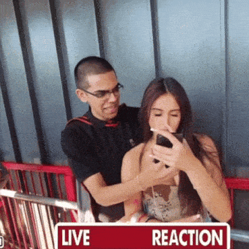 a man looks at a woman 's cell phone with a live reaction sign behind them