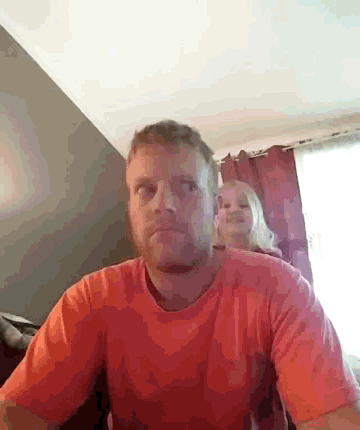 a man in an orange shirt is sitting in a room with a girl behind him .