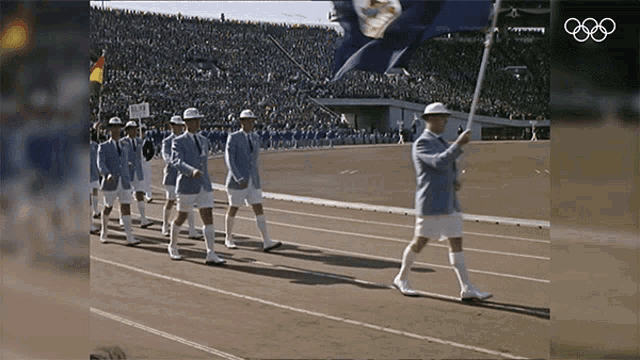 a group of people marching down a track with the olympics logo in the corner