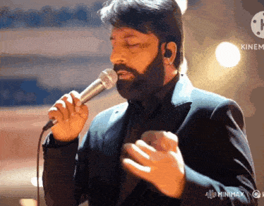 a man with a beard is singing into a microphone with a kinemax logo in the background