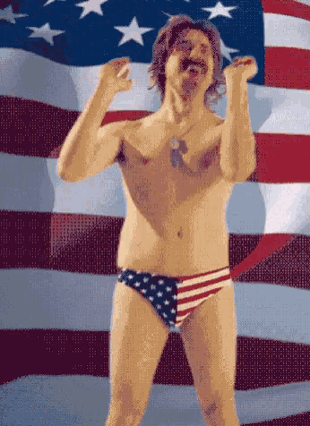 a shirtless man in an american flag swim trunks stands in front of a waving american flag