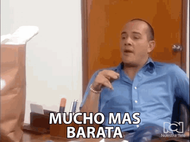 a man in a blue shirt is sitting at a desk with the words mucho mas barata above him