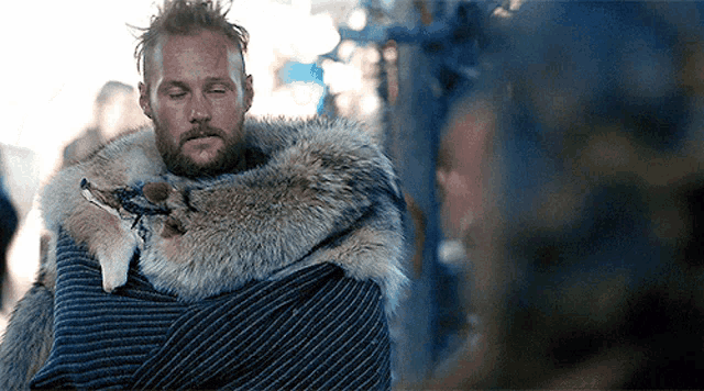 a man with a beard is wrapped in a fur coat holding a fox