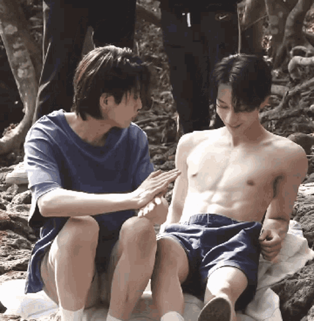 a shirtless man sits next to another shirtless man on a blanket