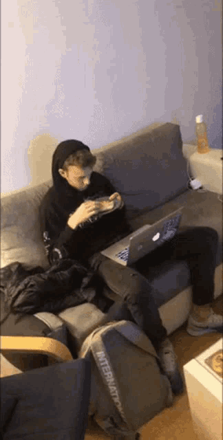 a man sits on a couch with a laptop and a backpack that says international