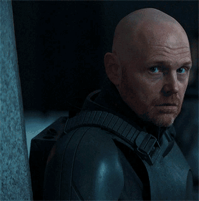 a bald man with a beard leans against a wall in a dark room