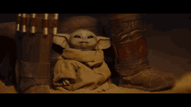 a baby yoda is sitting in the sand next to a person wearing boots