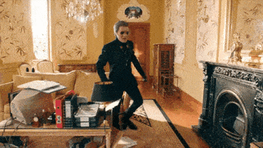 a man in a black suit is standing in a living room with a fireplace