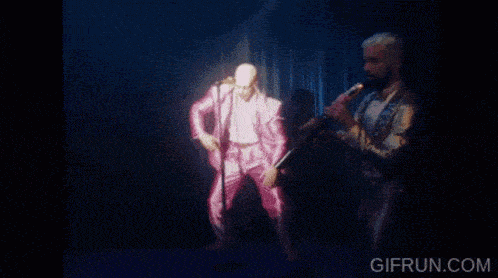 a bald man in a pink jacket is singing into a microphone on stage .