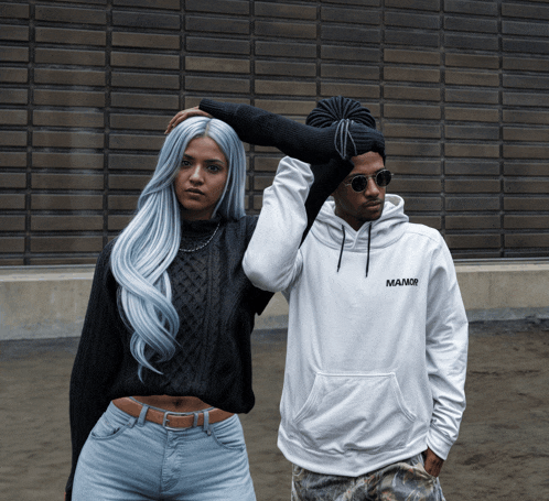a man wearing a white manor hoodie stands next to a woman in a black sweater
