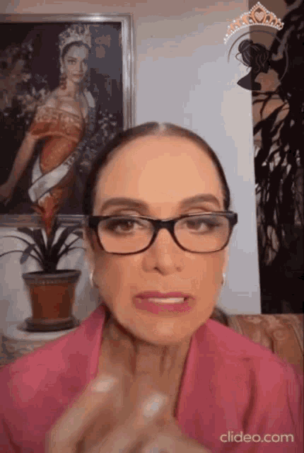 a woman wearing glasses is sitting in front of a framed picture of a woman in a crown .
