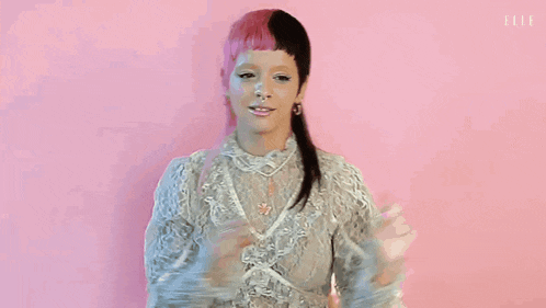 a woman with pink and black hair is standing in front of a pink background wearing a white lace dress .