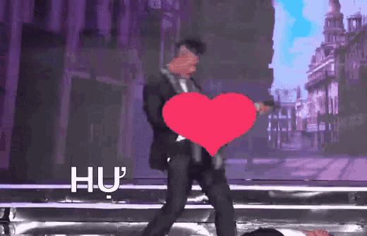 a man in a suit is holding a pink heart in front of his face and hu is written on the screen behind him