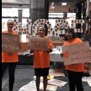 three women in orange shirts are holding up signs one of which says " hope "