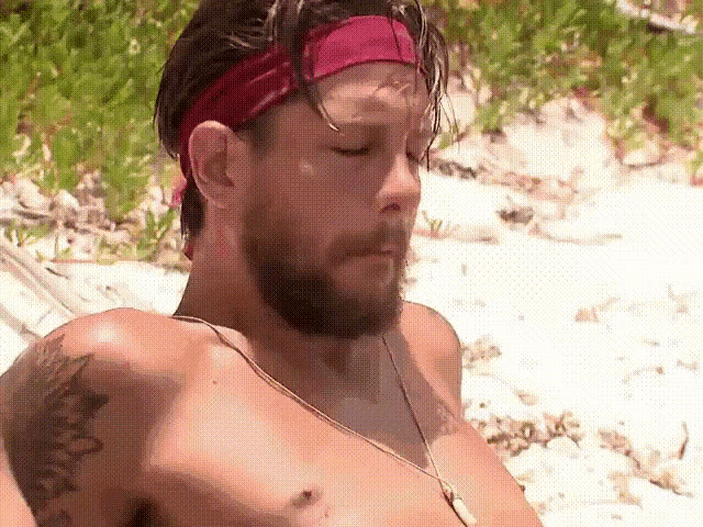 a shirtless man with a beard and a red headband is sitting on a beach .