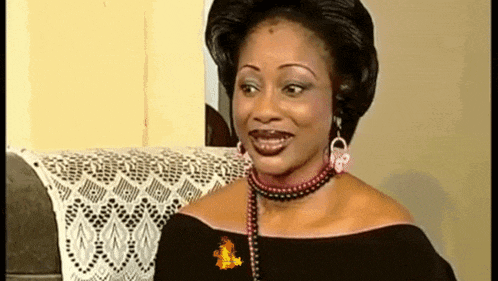 a woman wearing a black dress and a pearl necklace is smiling while sitting on a couch .