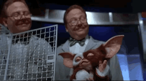 a man is holding a stuffed animal while another man holds a cage with a gremlin in it .