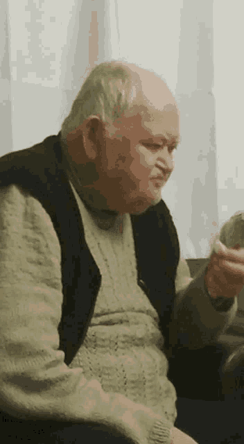 an elderly man wearing a green sweater and a black vest is holding something in his hand