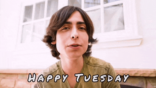 a young man in a green shirt says happy tuesday in front of a window .