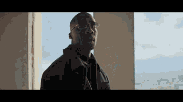 a man wearing glasses and a black jacket is standing in front of a window looking out .