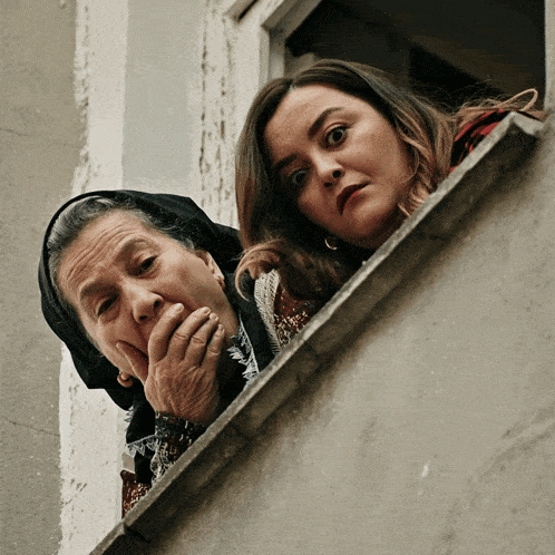 two women are looking out of a window and one is covering her mouth with her hand