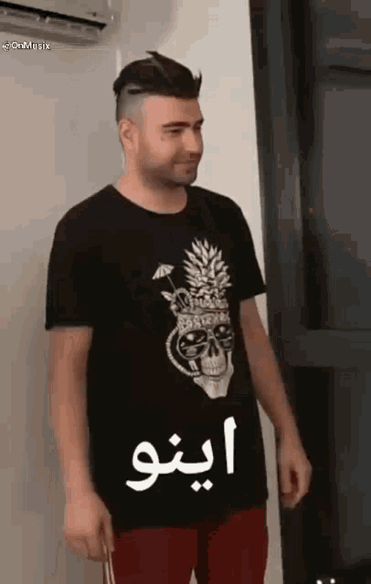 a man wearing a black t-shirt with a skull and pineapple on it is standing in front of an air conditioner .