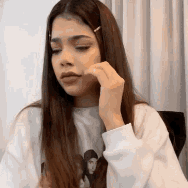 a woman with long brown hair is wearing a white sweater and holding her hand to her chin .