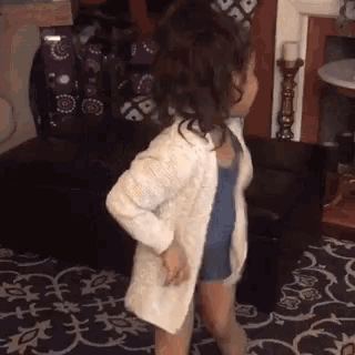 a little girl is dancing in a living room while wearing a white sweater and overalls .