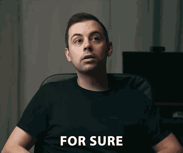 a man in a black shirt is sitting in a chair with the words for sure written on his shirt