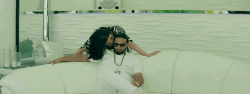 a man and a woman are sitting on a couch and the man has a necklace with the letter t on it