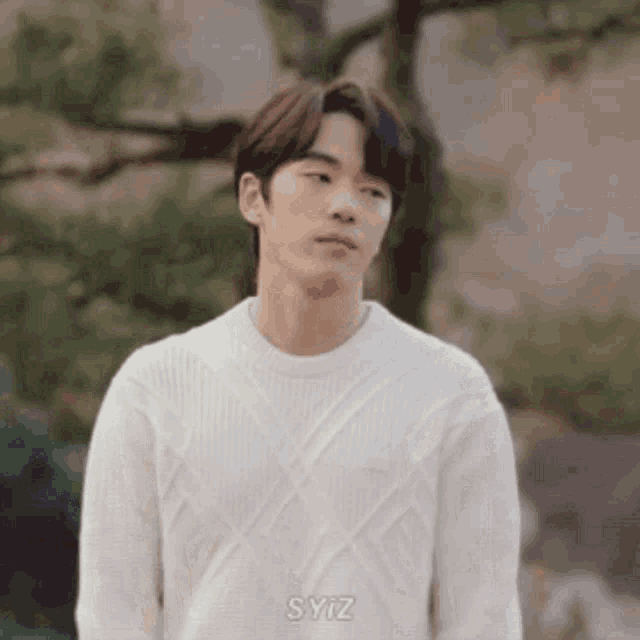 a young man in a white sweater is standing in front of a tree and looking at the camera .