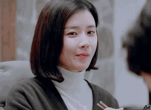 a woman wearing a white turtleneck and a black cardigan holds chopsticks in her hand