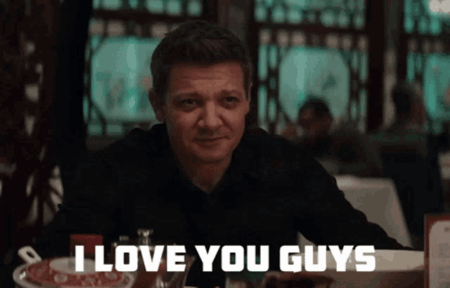 a man sitting at a table with a sign that says i love you guys on it