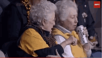 two elderly women are sitting in a stadium watching a game and one is holding a can of rootbeer .