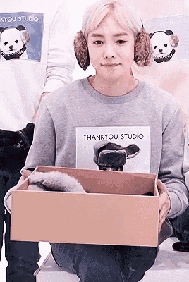 a woman wearing ear muffs and a thankyou studio sweatshirt is holding a cardboard box .