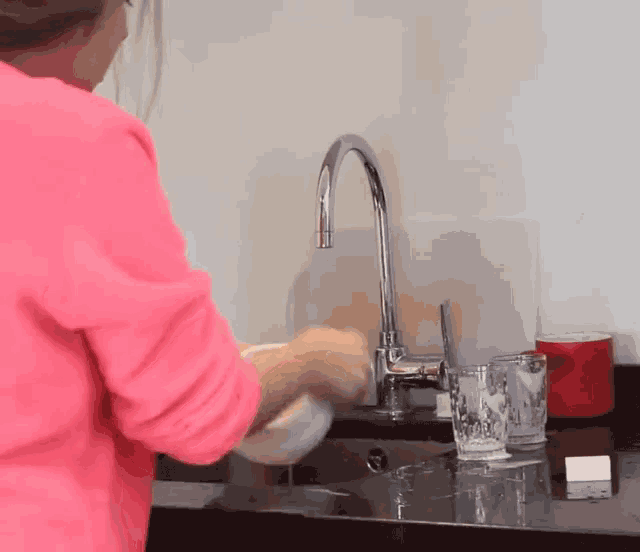 a woman in a pink shirt washing dishes in a kitchen sink